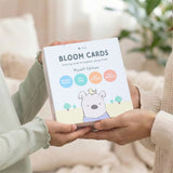 Two hands holding a pack of 'Bloom Cards' with a bear illustration on a blurred indoor background.